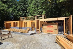 Exterior view of a residential construction site with wooden framing, showcasing the structure of walls and roof under a clear sky.