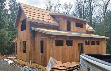 Construction site of a house with partially completed wooden framing, showing exposed walls and roof structure, with truck tailgate in the foreground.
